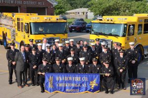 A group of firefighters in formal uniforms pose outdoors in front of two yellow fire trucks, holding a blue banner reading "Hy-View Fire Co. Cheektowaga, N.Y." on a sunny day.