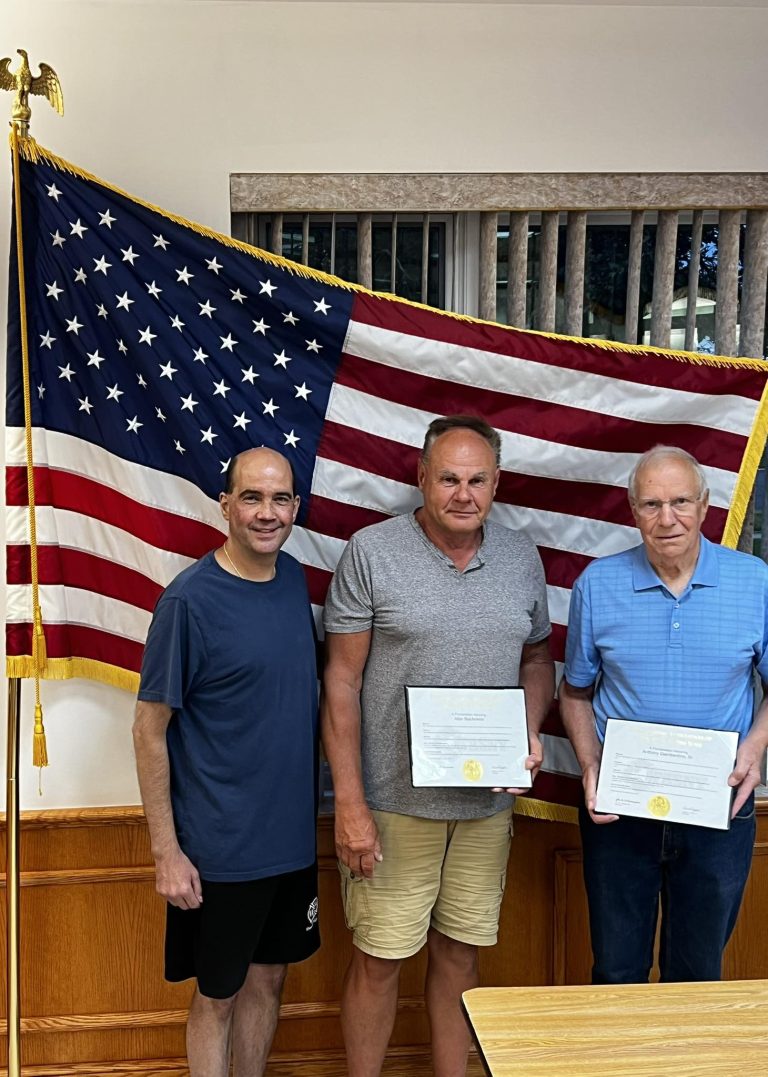 Three men stand indoors in front of a large American flag. Two of them hold certificates, while the third stands on the left in casual clothes. They are posing and smiling for the photo.