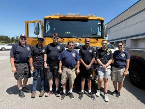 A group of seven people in matching fire department shirts stand in front of a yellow fire truck on a sunny day, smiling at the camera.