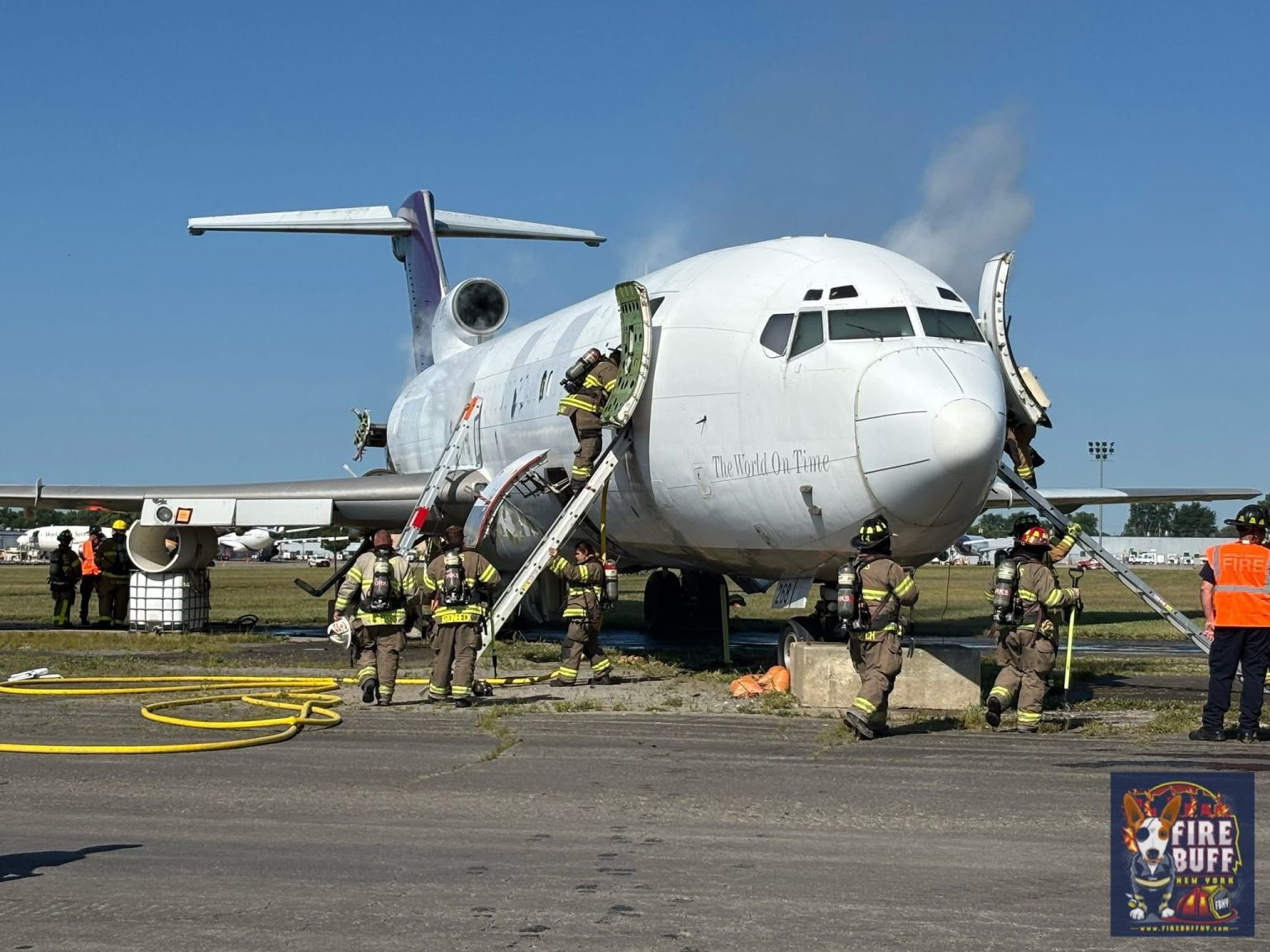 Firefighters in full gear respond to smoke coming from a white airplane on the ground, using hoses and equipment. A few people observe nearby, and a "Fire Buff" event logo is visible in the bottom right corner.