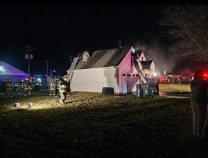 Firefighters work at night to extinguish a house fire. Smoke rises from the roof as firefighters use ladders to access the building. Emergency vehicles and a crowd are visible in the background.