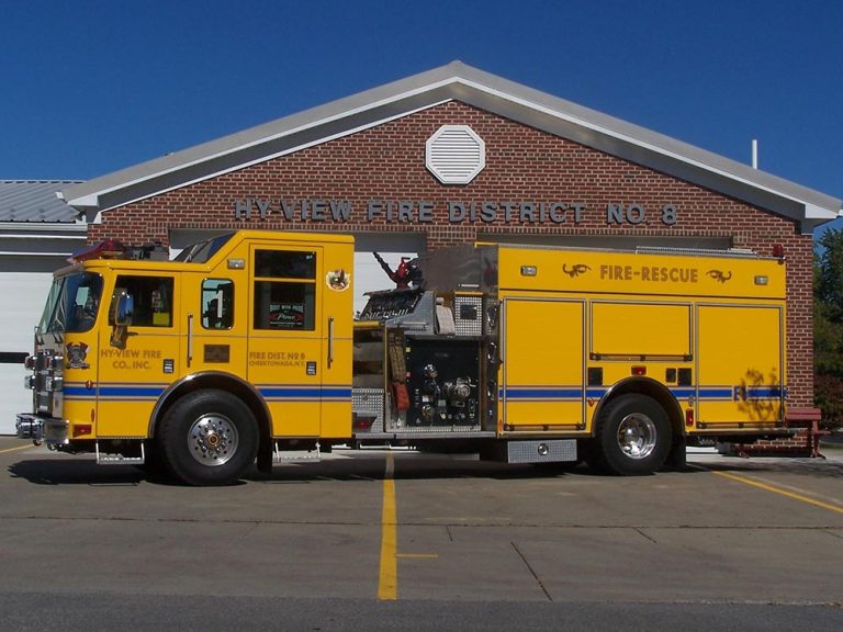 A yellow fire-rescue truck is parked in front of a brick building with a sign reading "HYVIEW FIRE DISTRICT NO. 8." The truck has multiple compartments and emergency equipment visible.