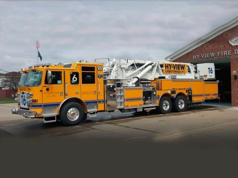 A yellow fire truck with blue and white accents is parked outside a brick fire station. The truck has a large ladder on top, and "HY-VIEW CHESTNUT AVE" is written on the side. The station sign reads "HY-VIEW FIRE DEPT.