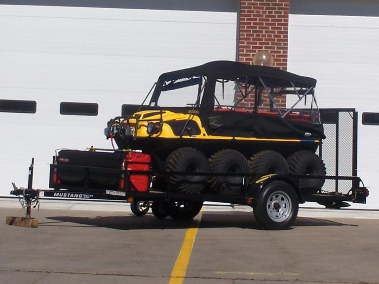 A yellow and black amphibious ATV with six wheels is parked on a trailer in front of a building with white garage doors and a brick column.