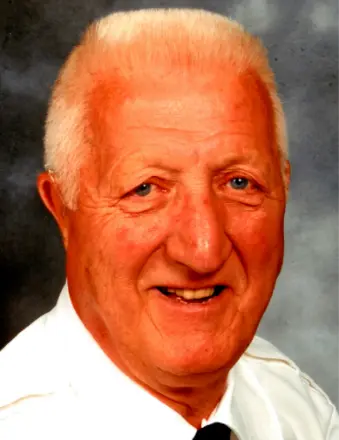 An older man with short, white hair is smiling. He is wearing a white collared shirt and a black tie, with a neutral, dark background behind him.