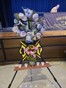 A heart-shaped floral wreath with pink and white flowers is displayed on an easel featuring a yellow firefighter emblem. Below are a vase of pink roses and six small lit candles. A man stands at a table in the background.