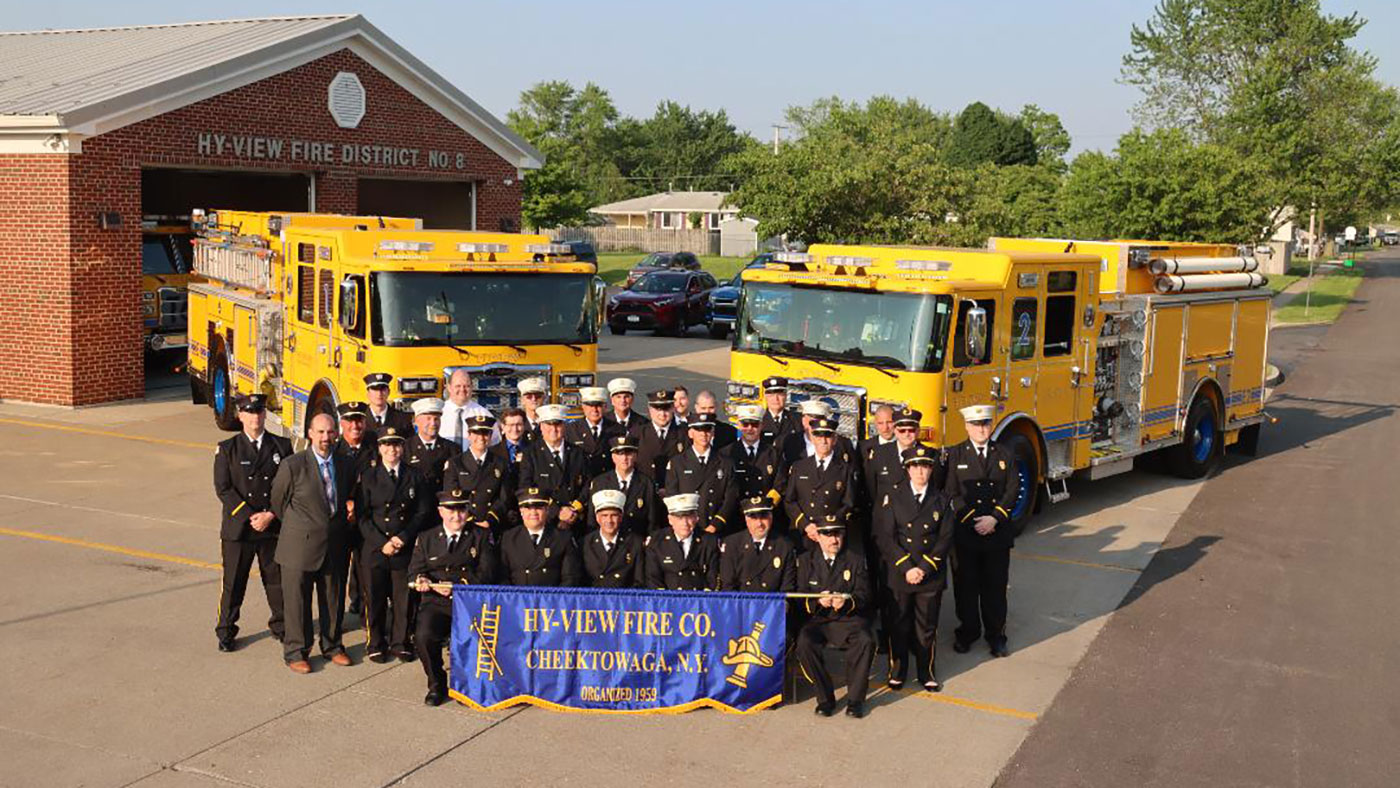 A group of firefighters in dress uniforms stands in front of two yellow fire trucks and a brick fire station, holding a blue banner that reads "Hy-View Fire Co. Cheektowaga, NY.
